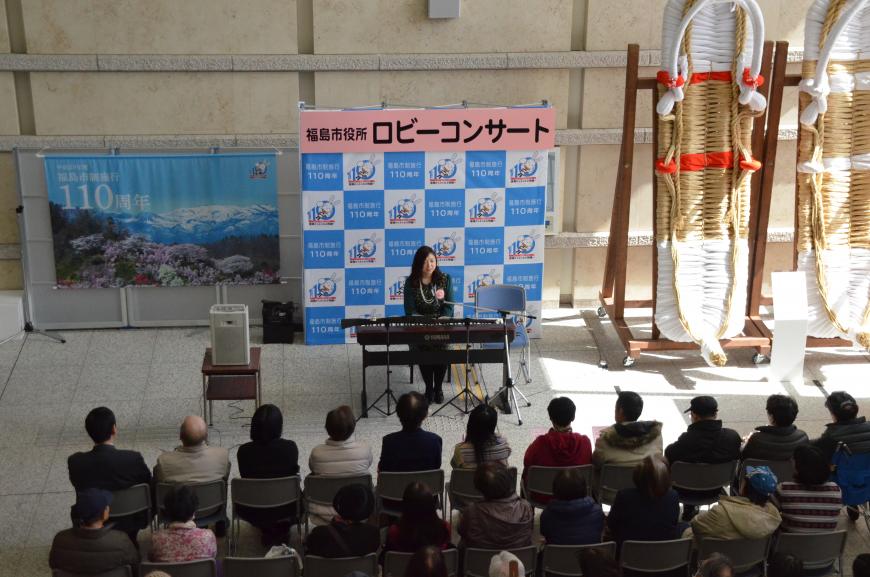 「福島市役所ロビーコンサート」と書かれたバックパネルの前に設置された電子ピアノの演奏席に星さんが、その前に置かれた客席の椅子にたくさんの来場者が座り、左側には110周年記念の垂れ幕、右側には大きなわらじが飾られている会場全体の写真