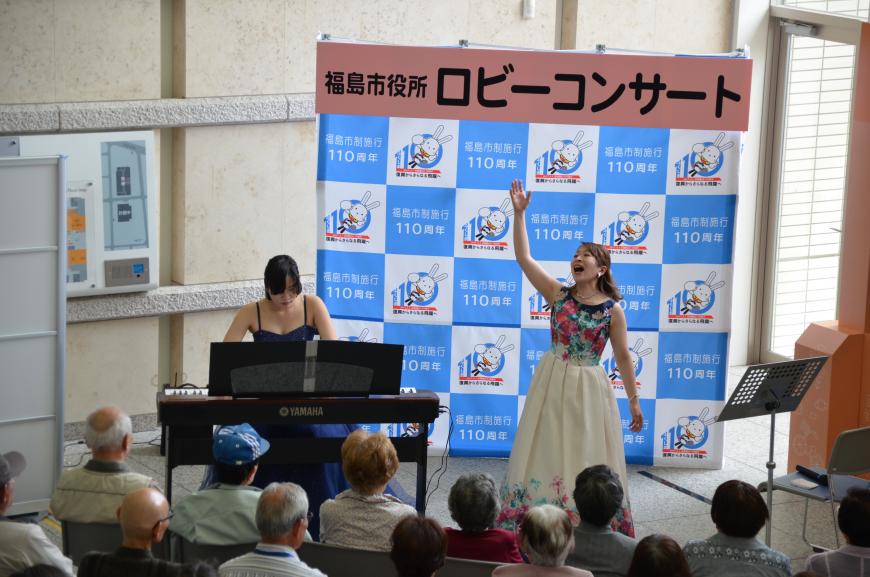 「福島市役所ロビーコンサート」と書かれたバックパネルの前で白地に花柄のドレスを着た女性が右手を上に伸ばしながら歌唱し、左側の青いドレスの女性が電子ピアノを演奏している写真