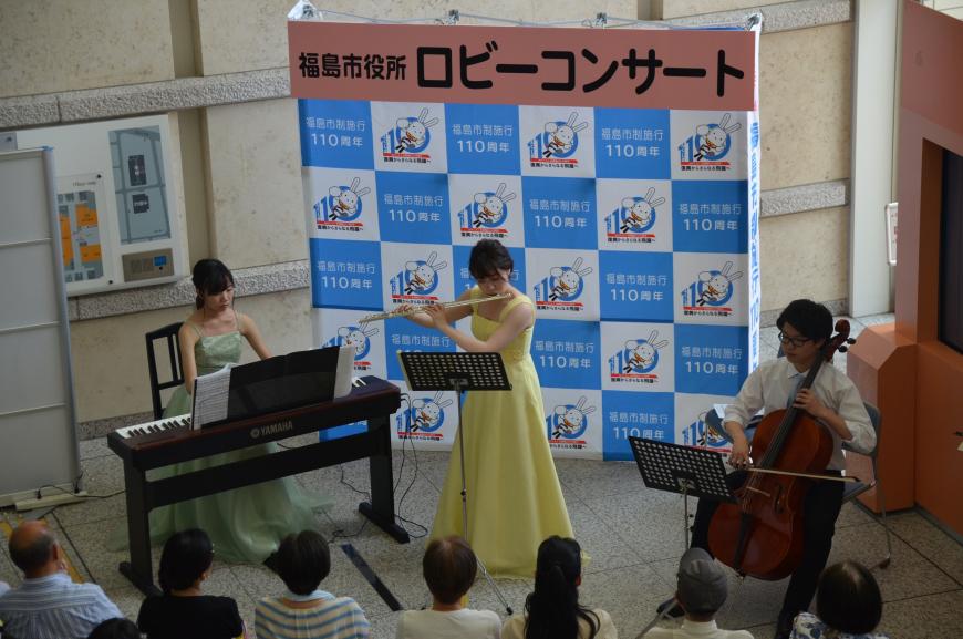 「福島市役所ロビーコンサート」と書かれたバックパネルの前で、薄緑のドレスを着た女性が電子ピアノ、黄色いドレスの女性がフルート、白いシャツの男性がチェロを演奏している写真