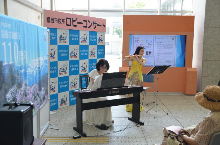 「福島市役所ロビーコンサート」と書かれたバックパネルの前で、手前の白いドレスの女性が電子ピアノを、奥の黄色いドレスの女性がフルートを演奏している写真