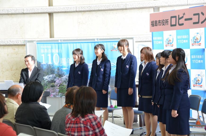 「福島市役所ロビーコンサート」と書かれたバックパネルと110周年記念の垂れ幕の前で紺色の制服を着た女子学生たちが並んで立ち、歌唱している写真