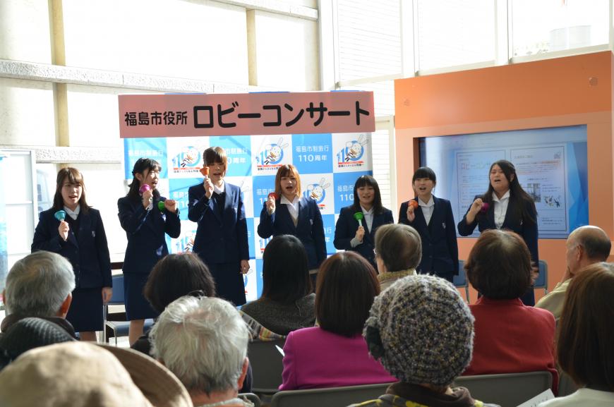 「福島市役所ロビーコンサート」と書かれたバックパネルの前で紺色の制服を着た女子学生たちが並んで立ち、ハンドベルの演奏をしながら歌唱している写真