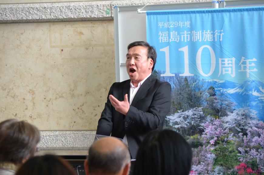 福島市制施行110周年記念の垂れ幕の前に立った黒いジャケットと白いシャツを着た男性が歌唱している写真