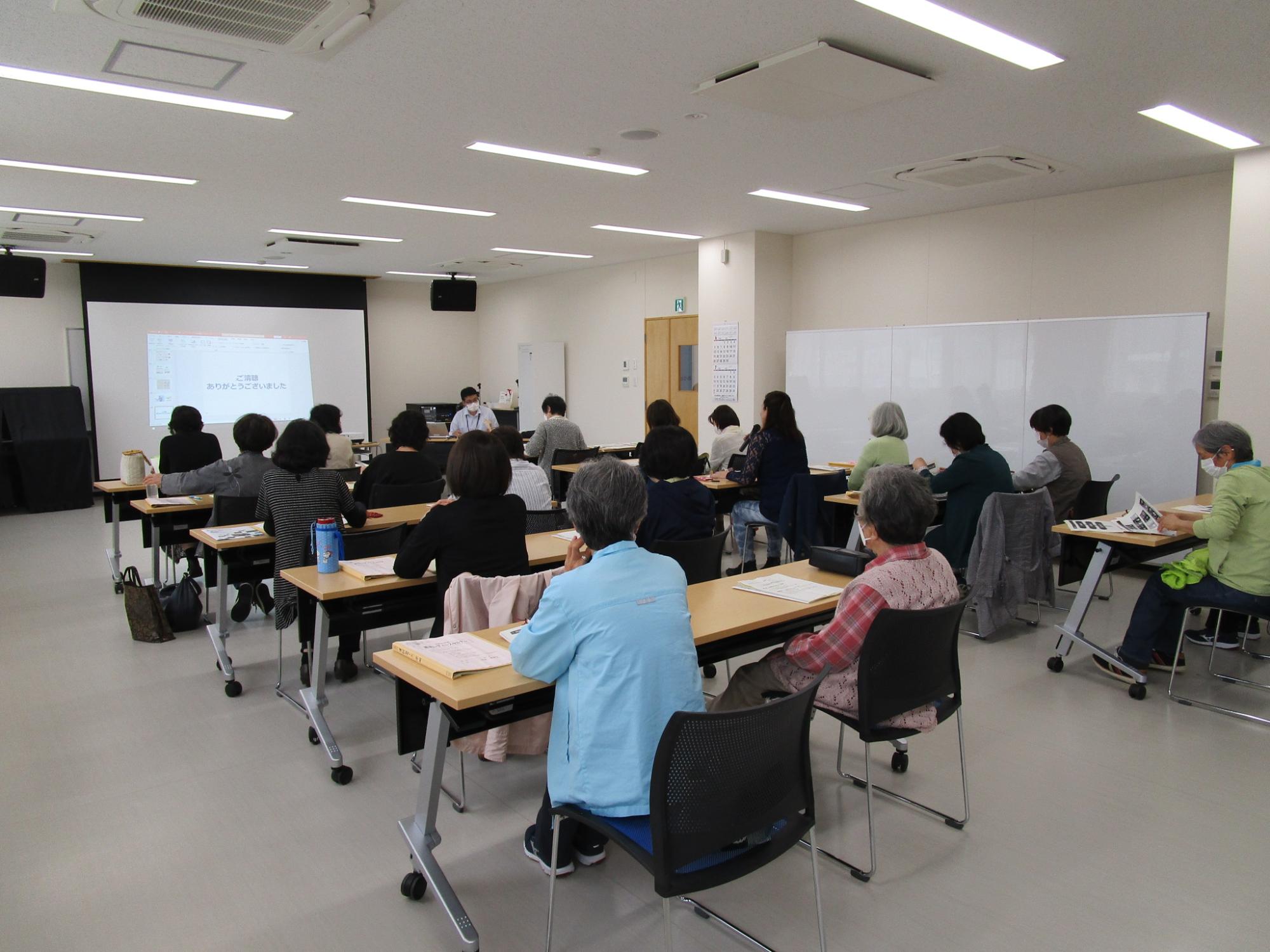大きなスクリーンの前に設置された長机に2名ずつ座り、3列目の女性がマイクで話をしている様子を会場の左斜め後方から写した写真