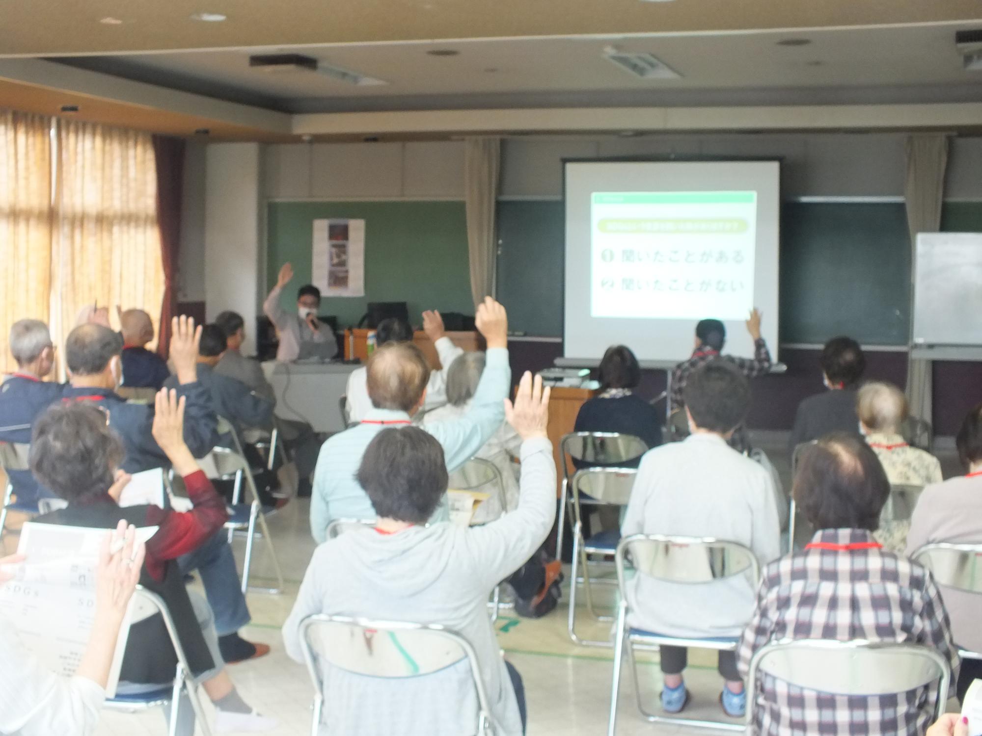 前方に大きなスクリーンがありその前のパイプ椅子に座っている参加者の人達数名が右手を挙手している様子を後方から写した写真