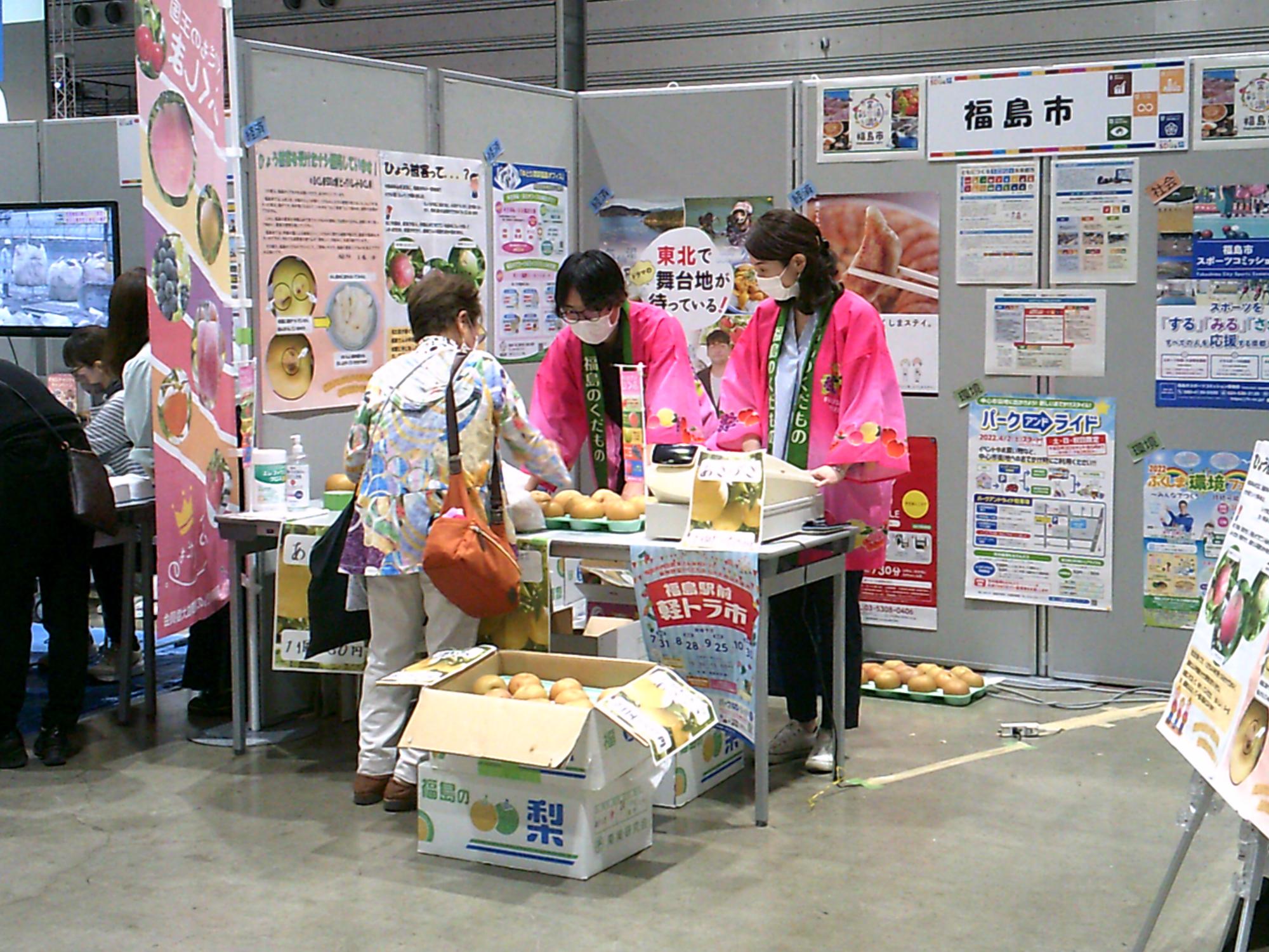 2名のピンクの法被を着た関係者の方が女性のお客様に商品の説明をしているふくしまのブースを写した写真