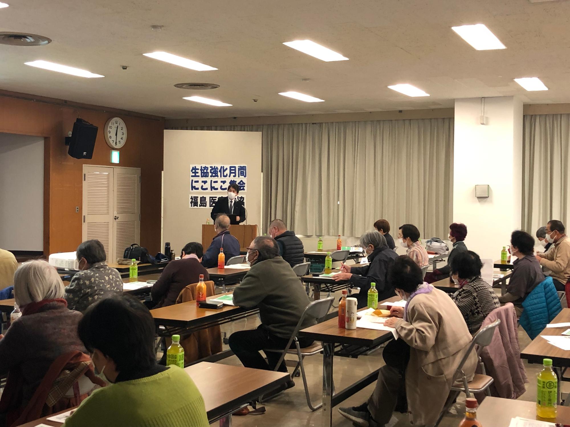 生協強化月間にこにこ集会と書かれた前に男性が立ち、その前の長机に参加者の人たちが座りながら説明を聞いている写真