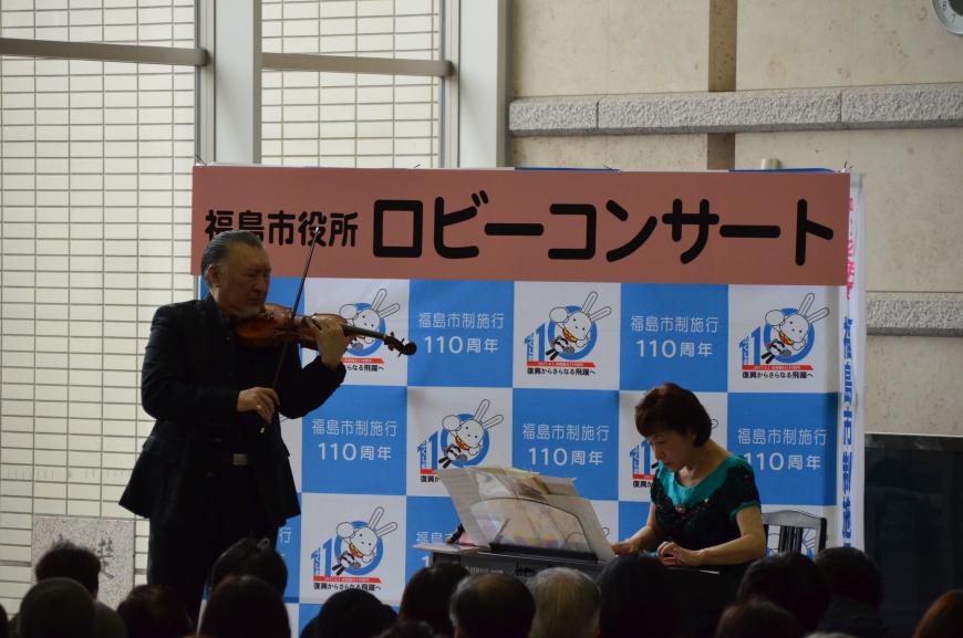 「福島市役所ロビーコンサート」と書かれたバックパネルの前で、女性が電子ピアノ、男性がヴァイオリンを演奏している写真