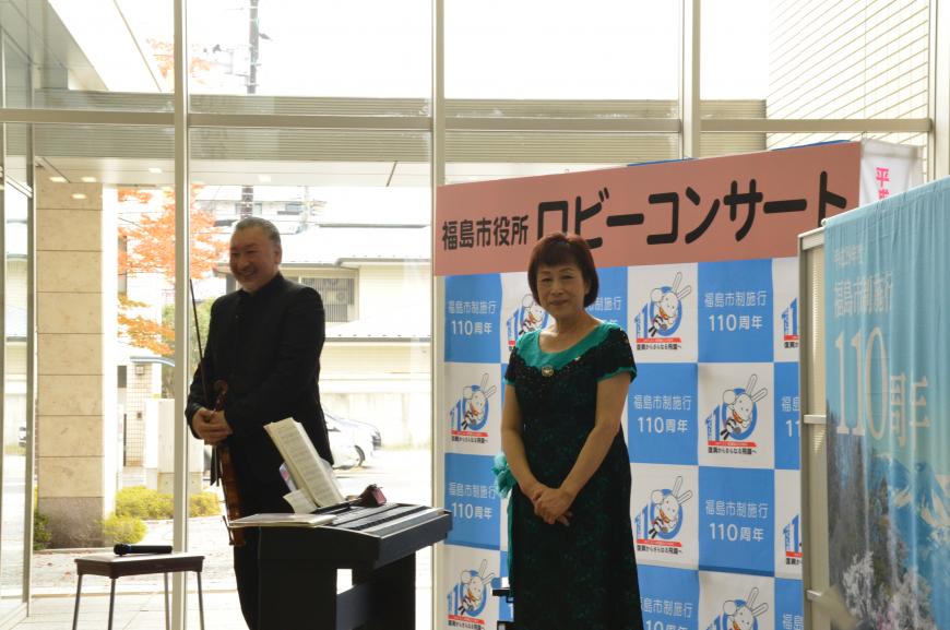 「福島市役所ロビーコンサート」と書かれたバックパネルの前に置かれた電子ピアノを挟み、男性と女性が笑顔で立って客席の方を見ている写真