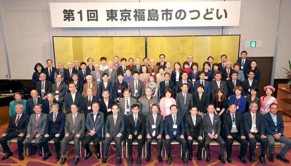 「第1回 東京福島市のつどい」と書かれた吊り看板が掲げられ、金屏風の前に5列ほどに並んだ参加者達の集合写真