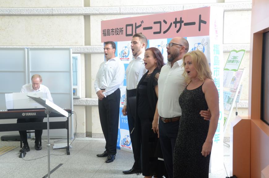 「福島市役所ロビーコンサート」と書かれたバックパネルの前で、白いシャツに黒いズボンの男性3名と黒いドレスの女性2名が歌唱し、左側で男性が電子ピアノを演奏している写真