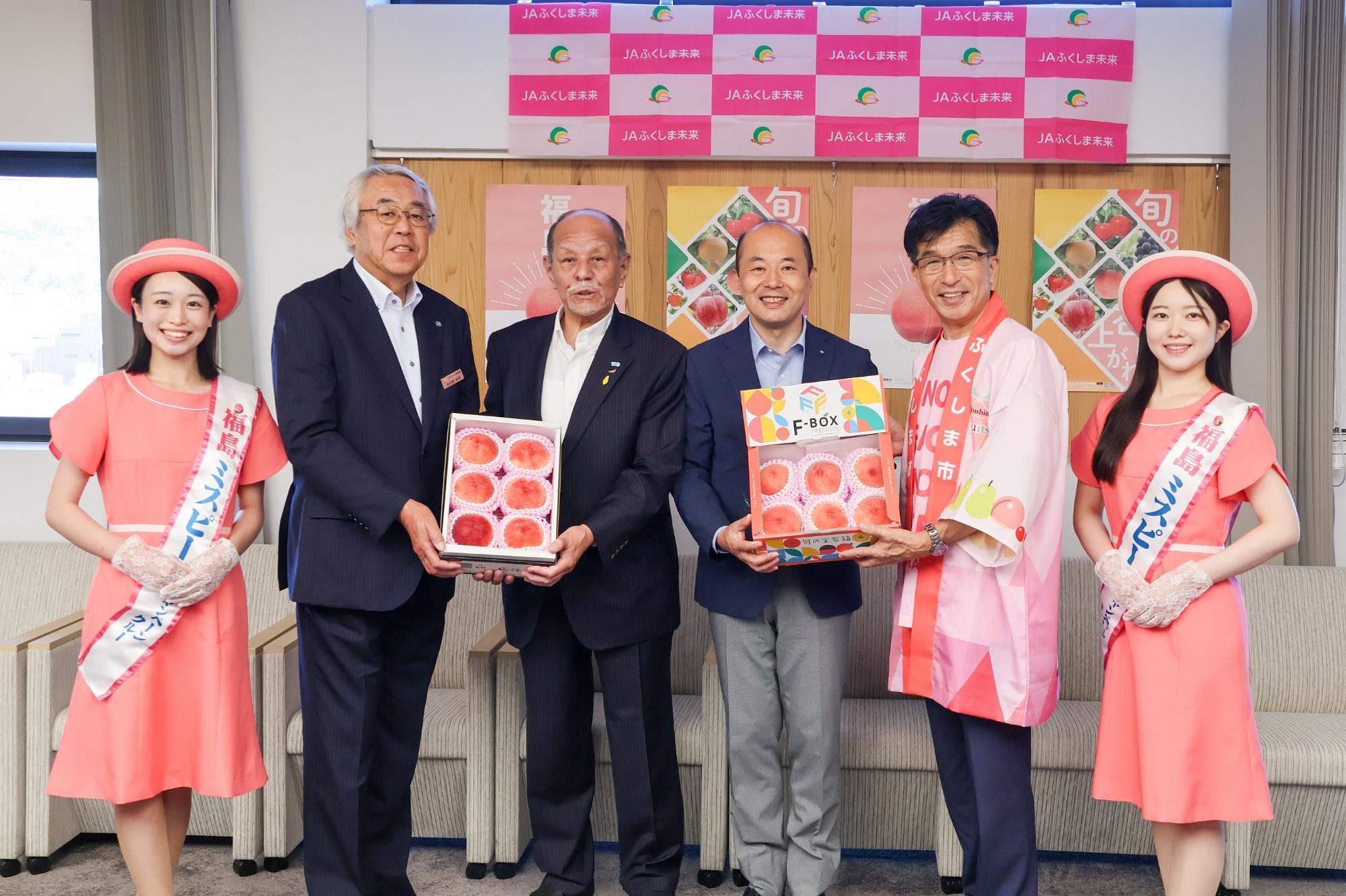 両側にミスピーチが立ち、その間に桃が入った箱を持っている市長と関係者の男性3名の写真