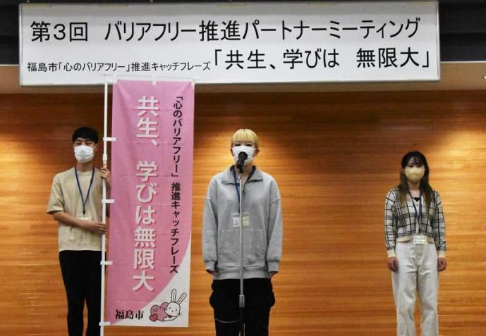 「共生、学びは無限大」と書かれたのぼり旗を持った関係者とマイクの前に立っている関係者、その隣に立っている女性の3名が舞台にたっている写真