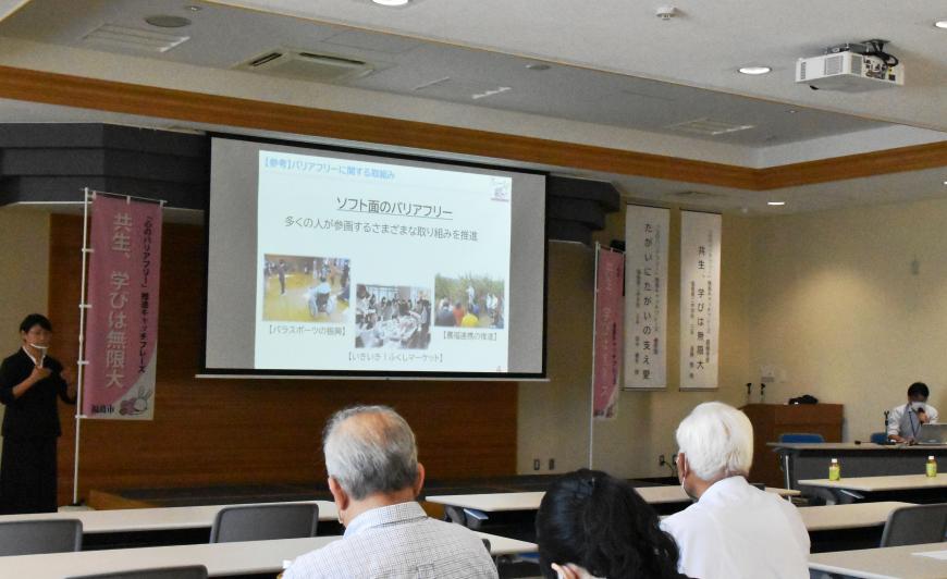 「共生、学びは無限大」と書かれたのぼり旗が両脇に設置され中央に資料が映し出されたスクリーンがある会場で参加者が話を聞いている様子を背後から撮影した写真