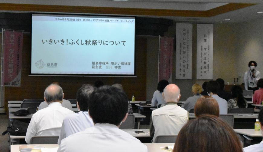 「いきいき!ふくし秋祭りについて」と書かれた資料がスクリーンに映し出されており、隣に設置されたテーブルで関係者がマイクを持って話をしている様子を参加者の背後から撮影した写真