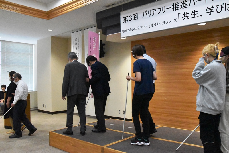 目隠しし、白杖を使用して歩く役と付添う役の二人一組で会場を一周するようすを撮影した「心のバリアフリー」出前講座のデモンストレーションの写真