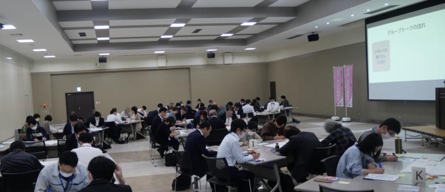 グループごとに分かれてそれぞれ長テーブルを組み合わせて数名の参加者が机を囲むように座り、資料に書き込んでいるグループトークの様子の写真