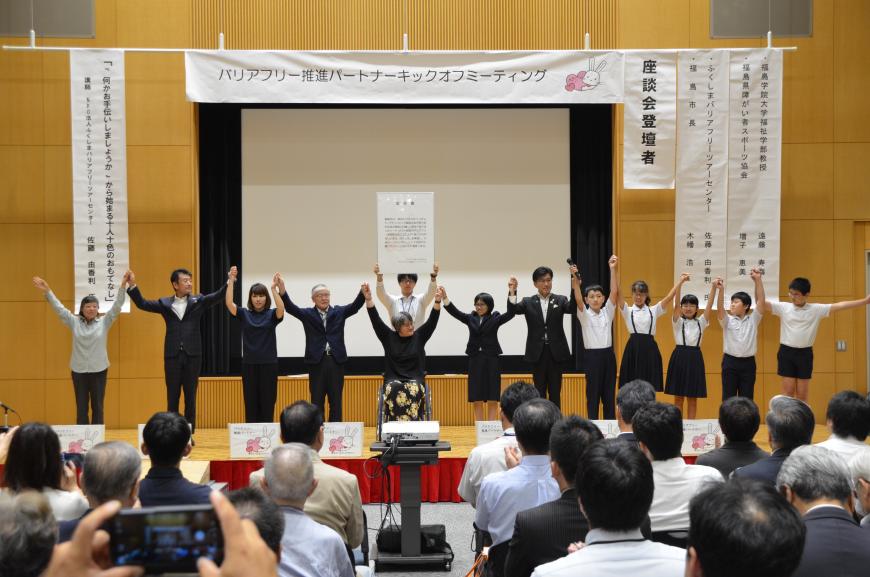 「バリアフリー推進パートナーキックオフミーティング」と書かれた横断幕を背景に車いすに乗った参加者を中心に市長や関係者、子どもたちが手を繋ぎ両手を頭上まで挙げているキックオフ宣言の様子の写真
