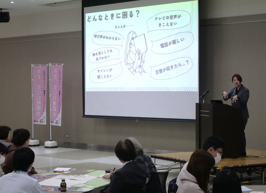 資料が映し出されたスクリーンの隣に演台が設置され、その前で手話で講演する講師の話に耳をかたむけている参加者たちの写真