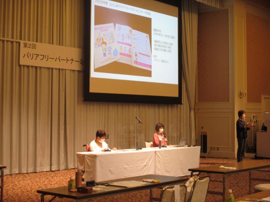 資料が映し出されたスクリーンの真下にテーブルが設置された席に座っている2名の関係者の写真