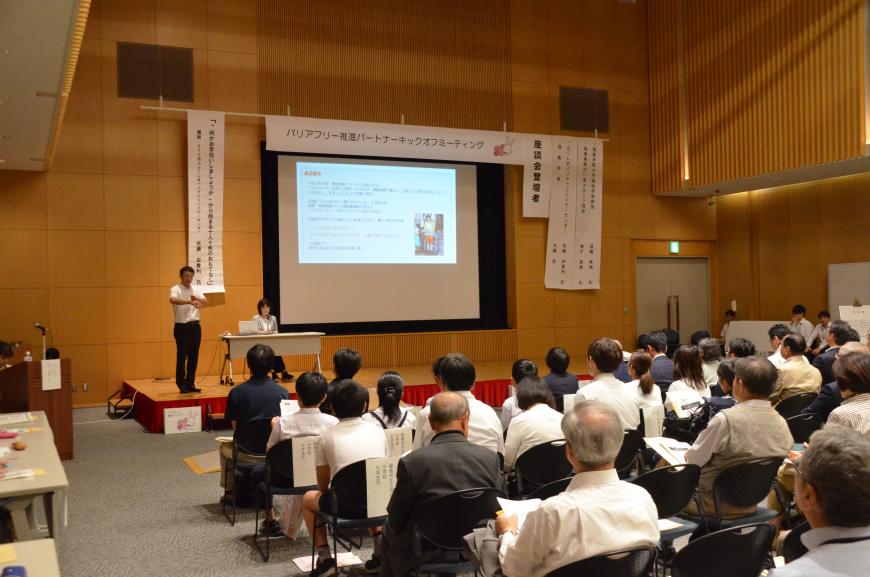 プロジェクターで資料が映し出されたモニターの横で関係者が話をしておりその様子を大勢の参加者が眺めている講演会の様子の写真