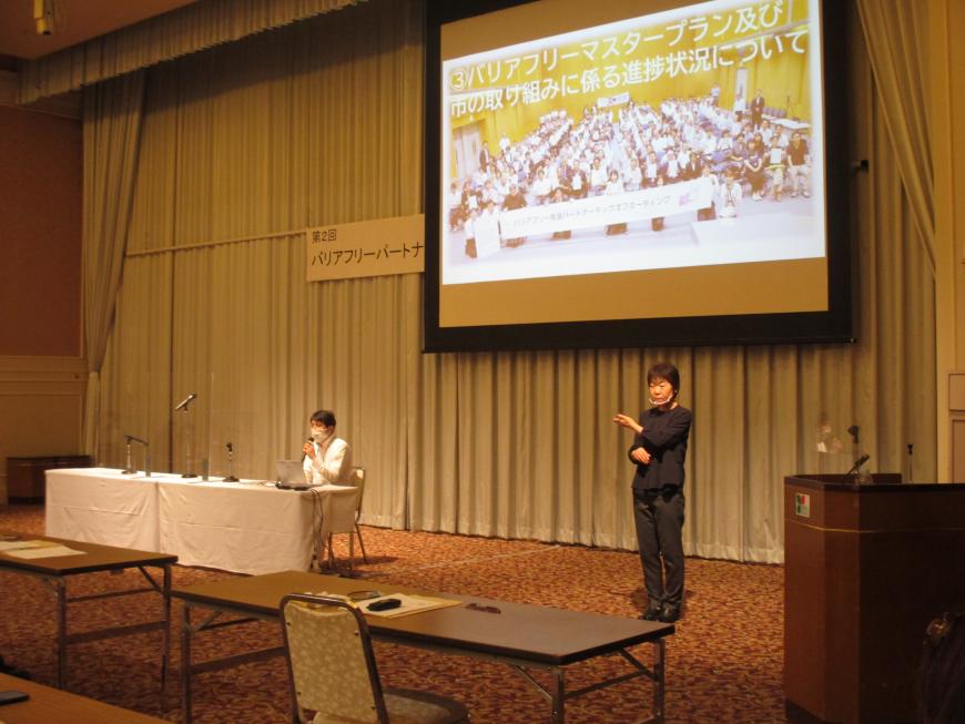 資料が映し出されたスクリーンの真下にテーブルが設置された席に座って話をしている関係者と、その傍で手話をしている女性の写真