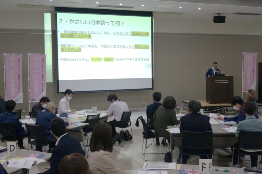 プロジェクターで資料を映し出したスクリーン画面を椅子に座っている参加者たちが眺めておりスクリーンの隣に設置された演台の前で講師が話をしているやさしい日本語セミナーの様子の写真
