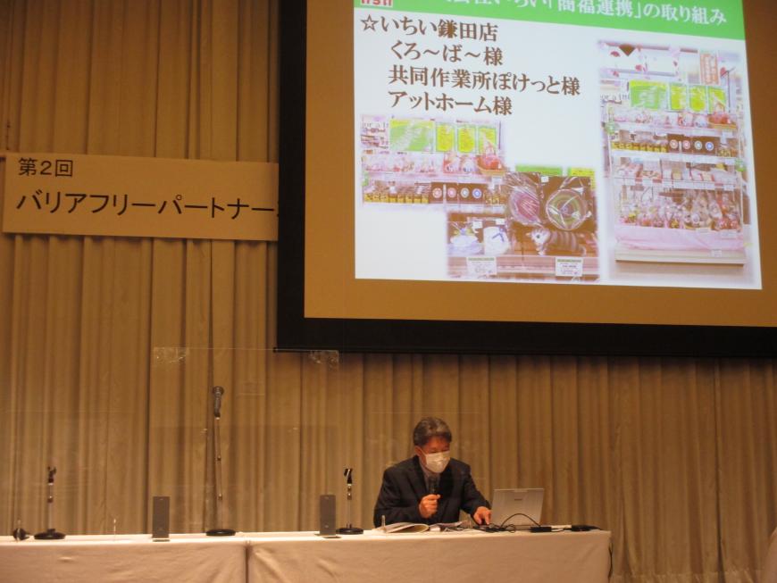 資料が映し出されたスクリーンの真下にテーブルが設置された席に座っている関係者の男性がマイクを持ち話をしている様子の写真
