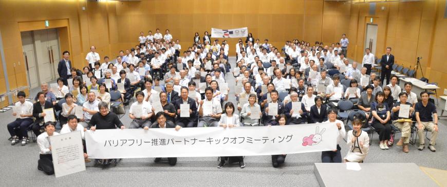 「バリアフリー推進パートナーキックオフミーティング」と書かれた横断幕を市長を含めた複数名が持ち、その後方で大勢の参加者たちがカメラ目線をしている記念撮影の写真