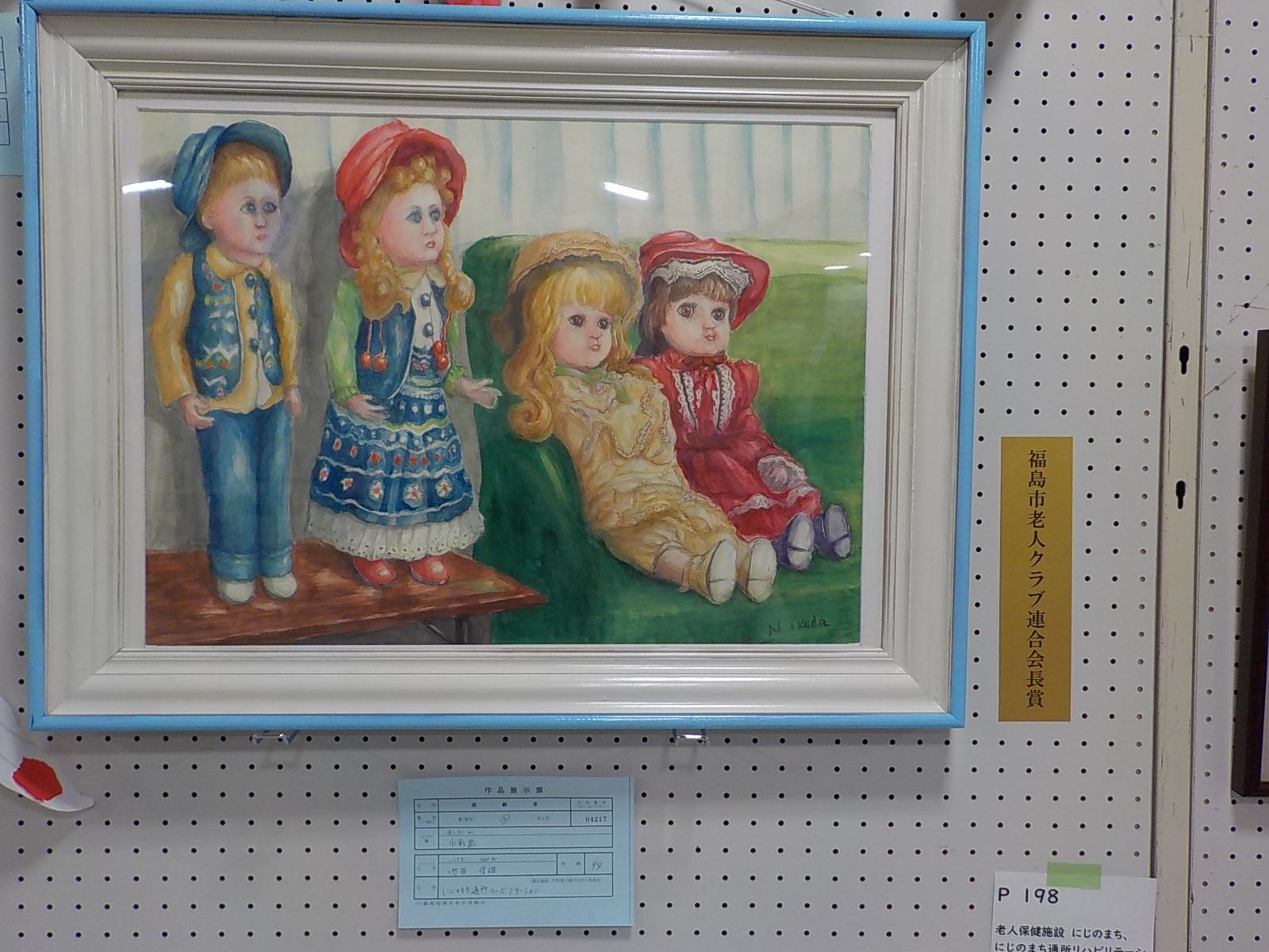 緑色のソファに黄色と赤色の服を着た2体の人形と、木製のテーブルの上に男の子と女の子の2体の人形が並んでいる様子を描いた福島市老人クラブ連合会長賞受賞作品の写真