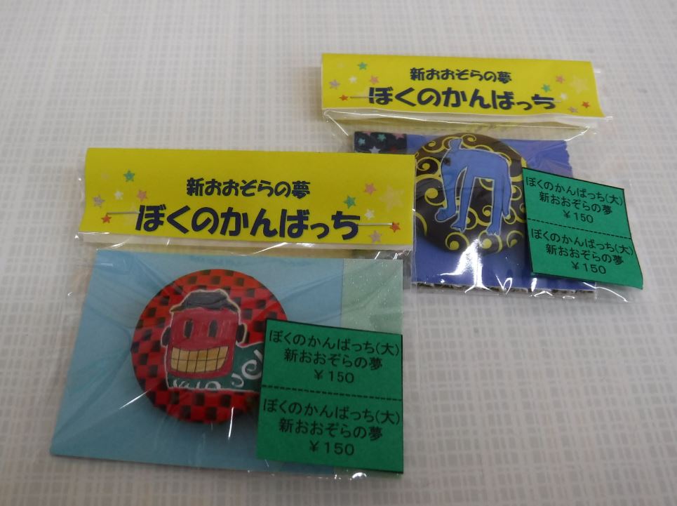 「新おおぞらの夢 ぼくの缶バッチ」と書かれた袋に2種類の缶バッチの商品が並んでいる写真