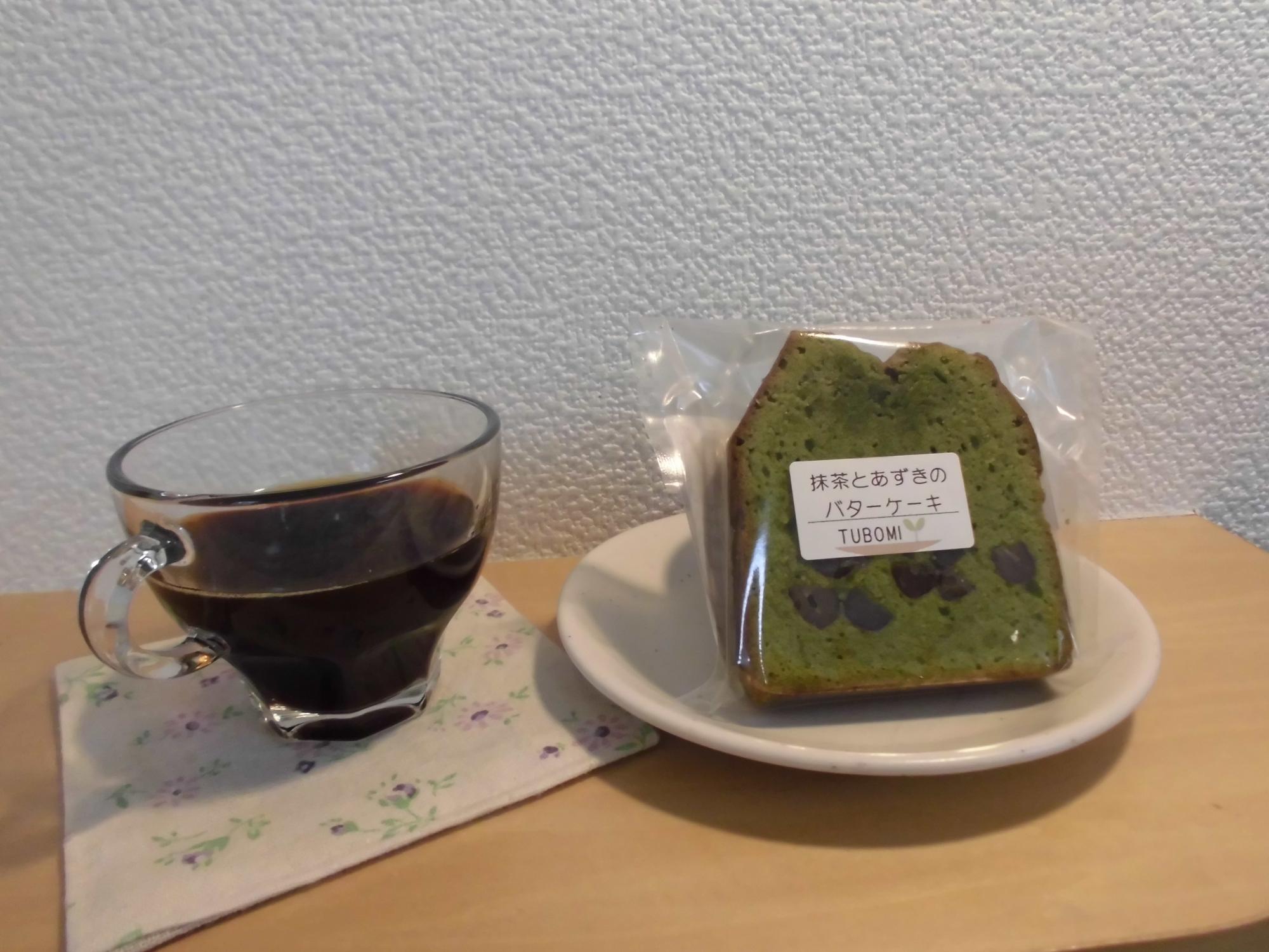 小花柄の布のコースターの上に置かれた透明のグラスに注がれた飲み物と、白皿の上に透明の袋に入った抹茶とあずきのバターケーキが置かれた写真