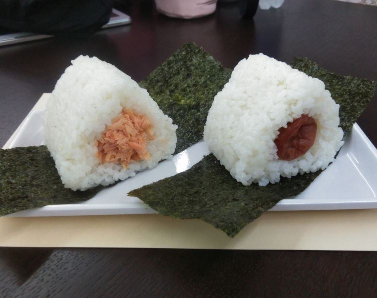 白色のトレーの上に海苔の上に鮭と梅干の具が中に入った2種類のおにぎりが置かれている写真