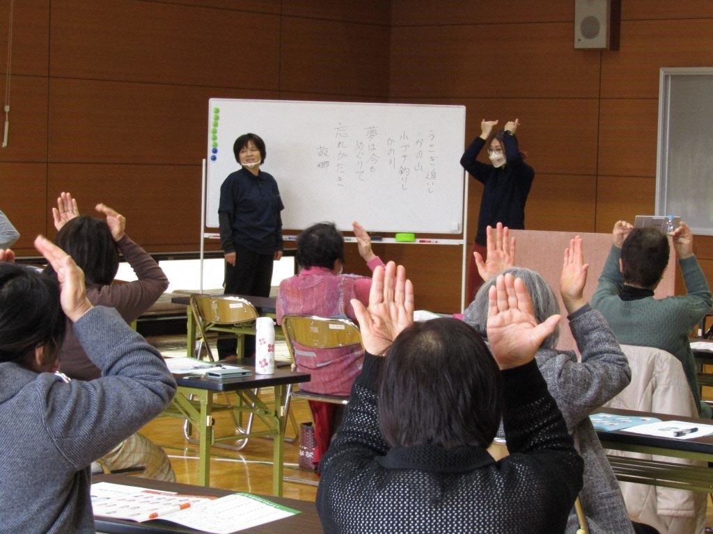 ホワイトボードの前に2人の女性講師が立ち、右側に立っている講師が両手を上に上げて手話を行っている様子を真似て、参加者が同じポーズをとっている講座の様子の写真