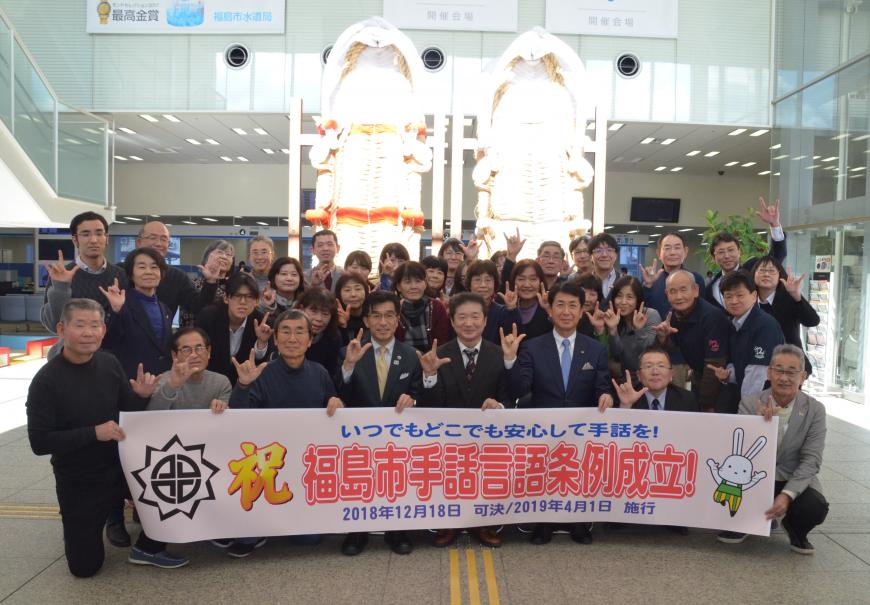「祝 福島市手話言語条例成立!」と書かれた横断幕を持った方々が後ろに並んでいる記念写真