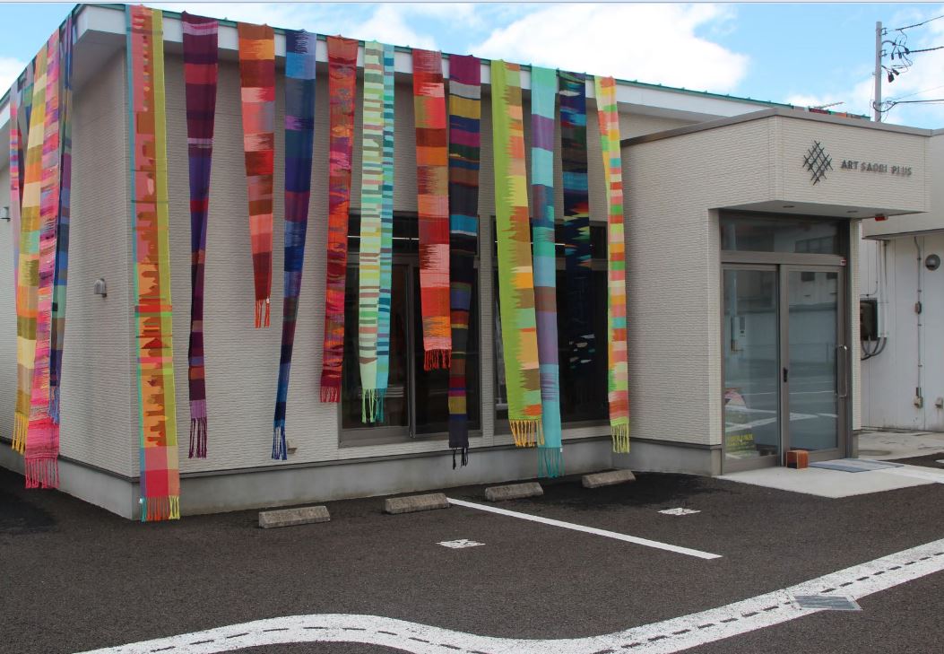 白壁の建物の中央の入り口から左半分の屋根から帯の様な形の色とりどりの作品が垂れ下がっている事業所の建物外観の写真