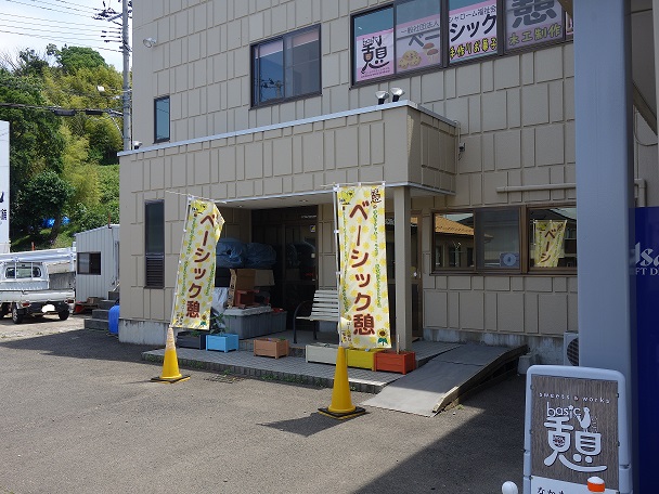 入り口前に「ベーシック憩」と書かれた2つののぼり旗が設置された、2階建てでベージュ色の外壁の建物外観の写真