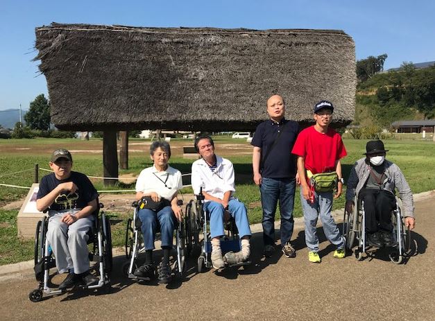 住居跡の建物をバックに、2名が立ち4名が車いすに座って並んでいる写真