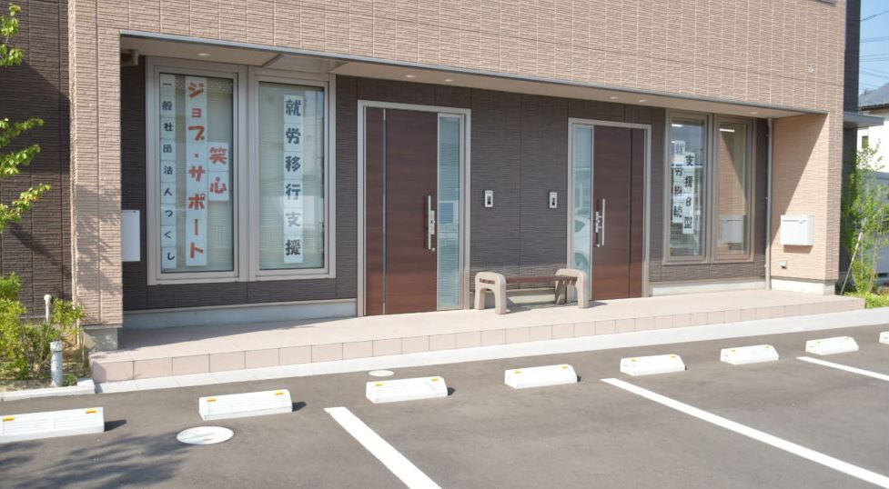 建物の前に駐車場が設けられ、窓ガラスに「就労移行支援」や事業所名が貼られているベージュ色の外壁のジョブ・サポート笑心の建物外観の写真
