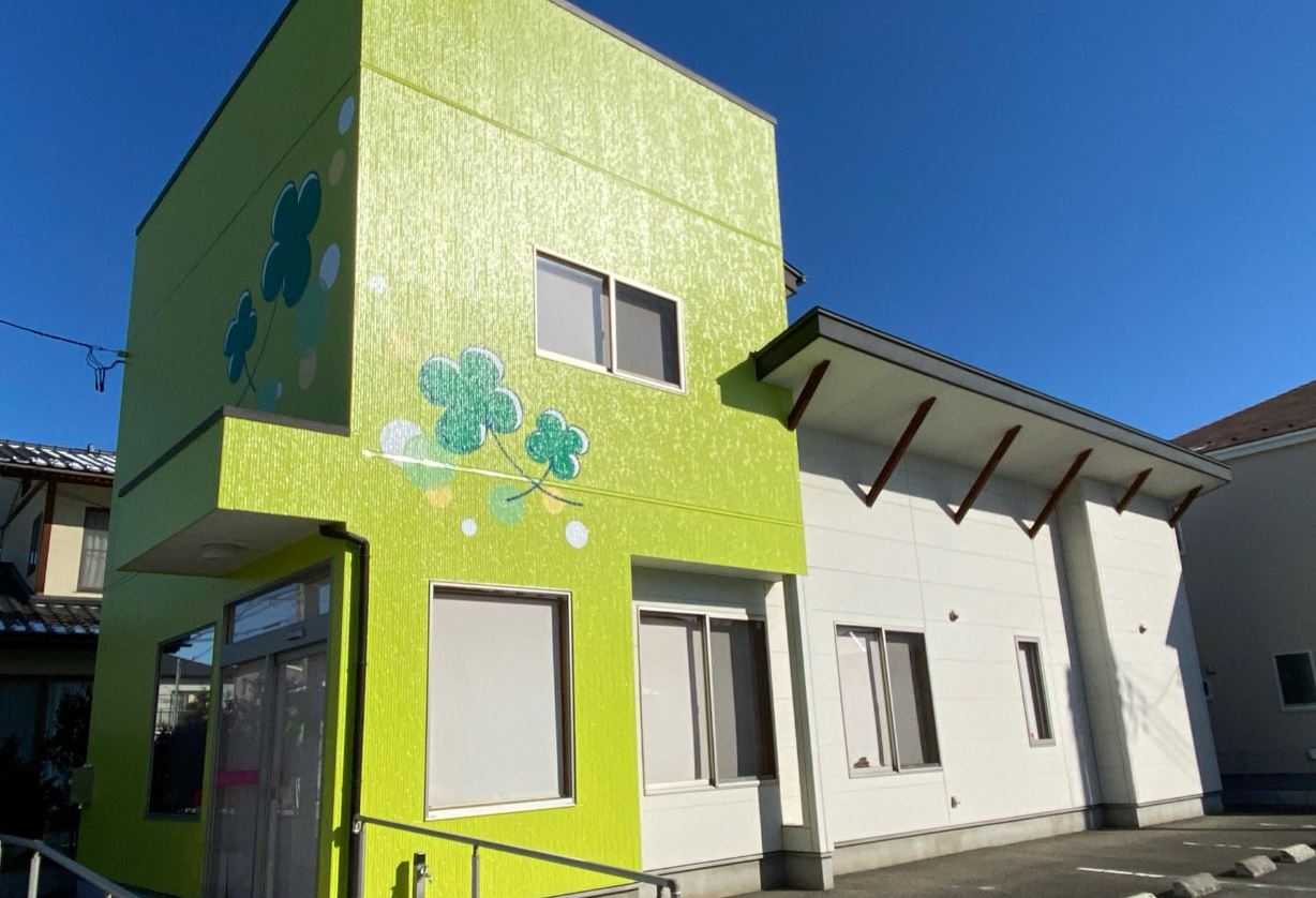 入り口前にスロープが設置され、右半分が1階の白壁で、左半分が2階建てで黄緑色の壁に大きな四つ葉のクローバーが描かれた建物外観の写真