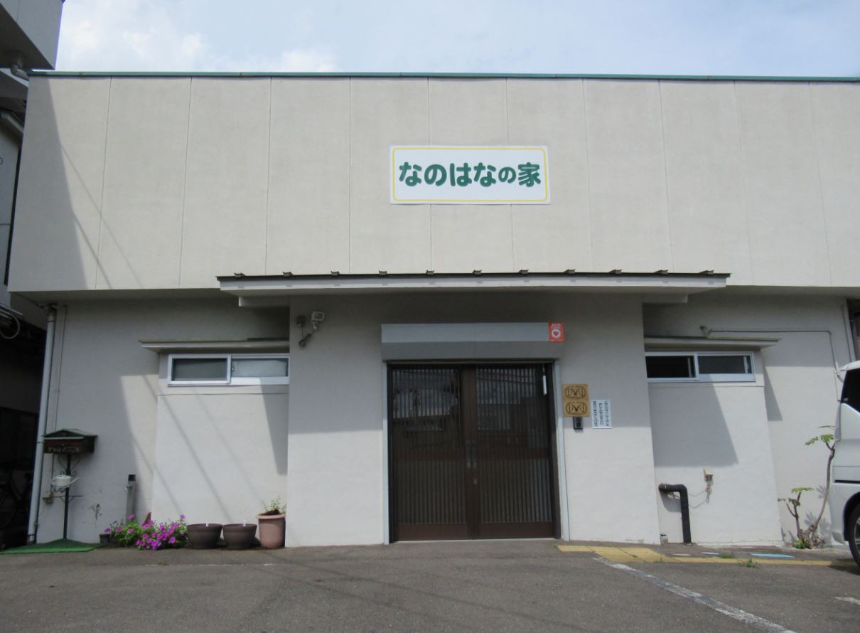 「なのはなの家」と書かれた看板が設置された白壁の建物外観の写真