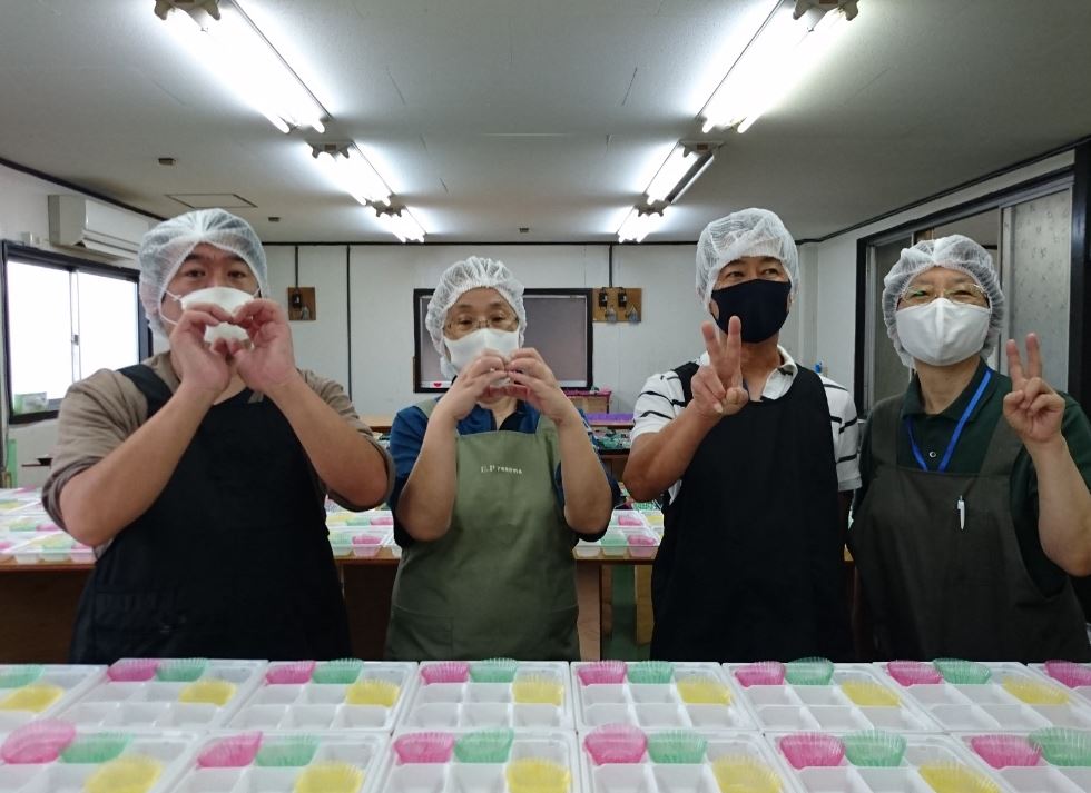 弁当容器が並んだ前でエプロンやマスクをつけた4人の利用者が横一列に並んでいる写真