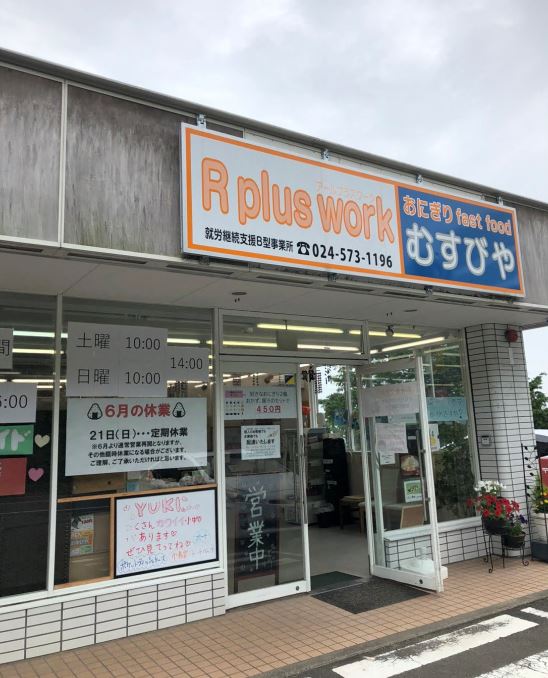 アルファベットで「アールプラスワーク」と書かれた看板が設置され、営業日時などのポップがガラス張りの窓に貼られ、入り口の右側の扉が開いた建物外観の写真