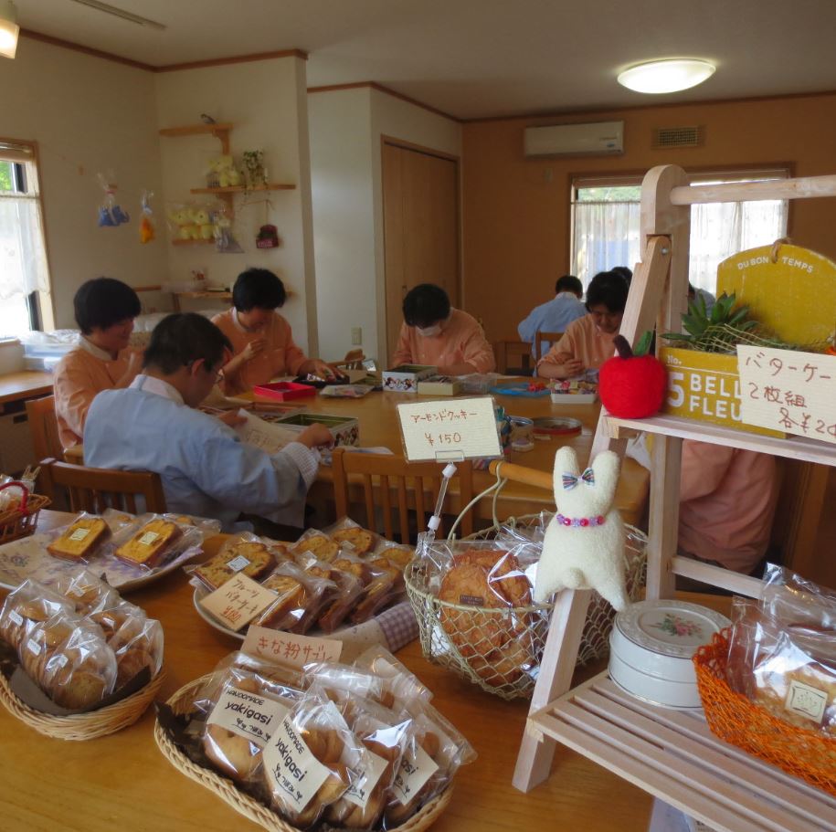 利用者の方々が作業を行っている手前に、種類ごとにカゴに入ったお菓子がテーブルの上に置かれた事業所内の写真