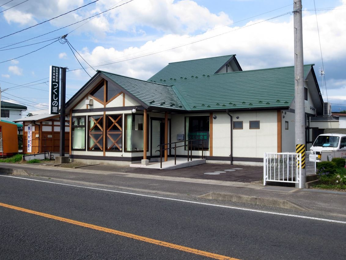 道路沿いに「つくしの里」と書かれた看板が設置された緑色の屋根で白壁の建物外観の写真