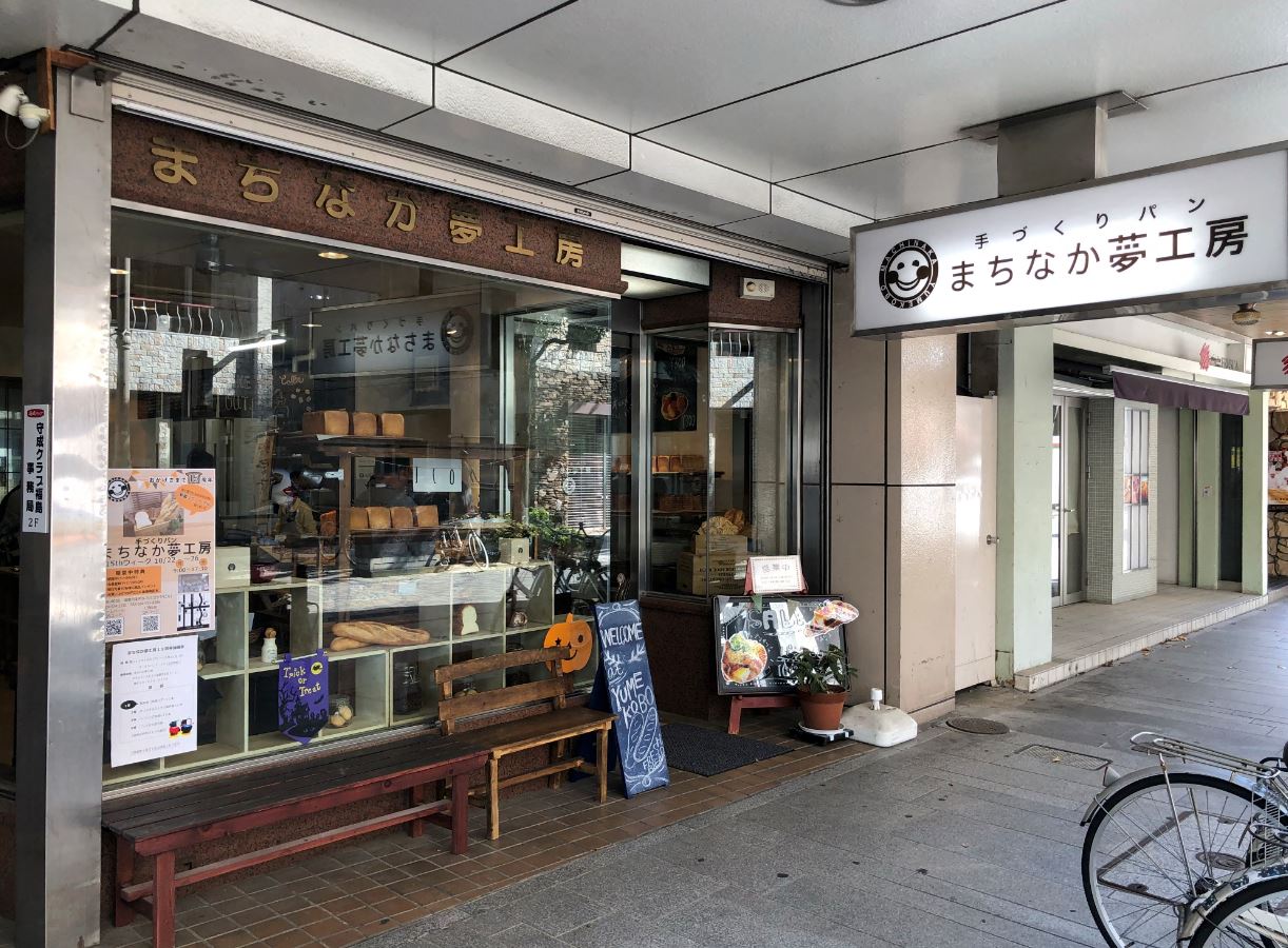 2台の木製ベンチが設置されたガラス張りの店舗入り口に「手作りパン まちなか夢工房」と書かれた看板が設置された写真