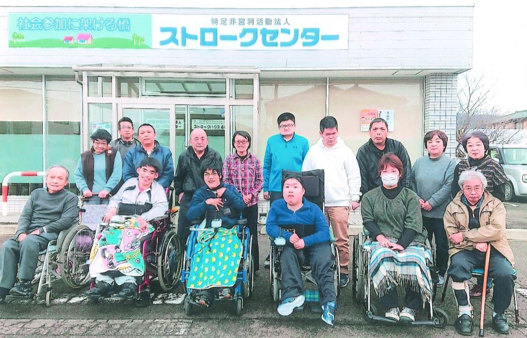 前列の椅子や車いすに6名、後列に10名の利用者が「特定非営利活動法人ストロークセンター」と書かれた事業所前に並んでいる集合写真