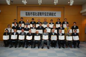 「福島市福祉避難所協定締結式」と書かれた横断幕が貼られた会場で、前列の椅子に10名後列に20名の参加者が立ち、協定書を開いてカメラに向かって見せている記念写真