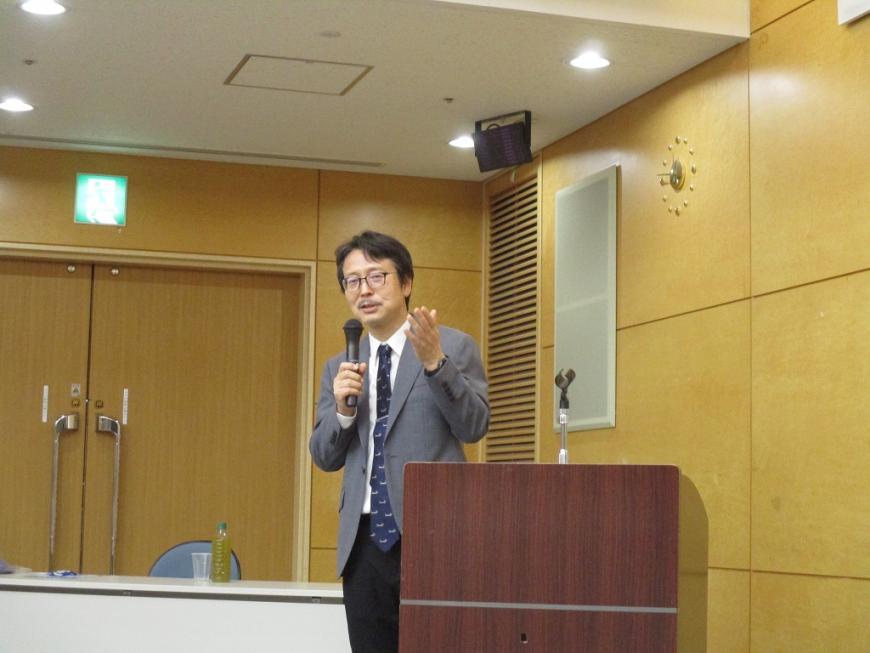 演台の横でマイクを持って立った男性が話をしている様子の写真