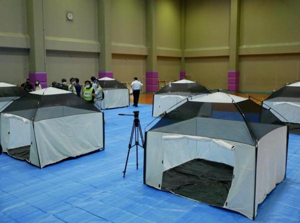 体育館の床にブルーシートが敷かれ、その上に屋根の一部がメッシュ素材になっている白いテントが複数並んでいる写真
